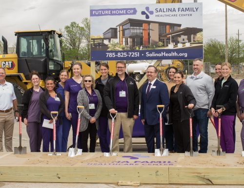 A crowd of people smile wearing suits or hospital scrubs hold shovels and stand on dirt in front of construction equipment