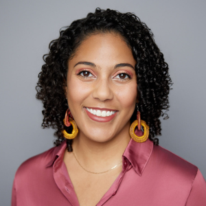 A woman with short black curly hair is facing the camera and smiling. She is wearing a pink top, colorful earrings, and a gold necklace. The background is gray. - Lauren Bowman