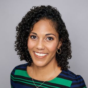 A woman with short curly black hair is facing the camera and smiling. She is wearing a green and blue striped shirt, gold hoop earrings, and a necklace. The background is gray. - Lauren Bowman