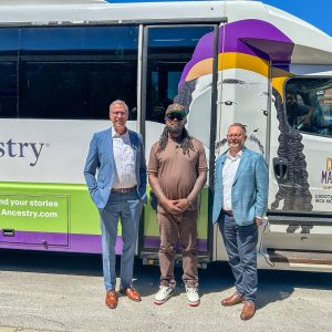 Three men stand in front of a bus decorated with a mural. They are smiling on a sunny day, conveying a friendly, celebratory mood.