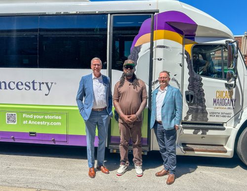 Three men stand in front of a bus decorated with a mural. They are smiling on a sunny day, conveying a friendly, celebratory mood.