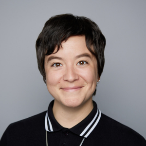 A Latine person with short brown hair is wearing a black top and necklace is facing the camera and smiling. The background is gray. - Carmen Aiken