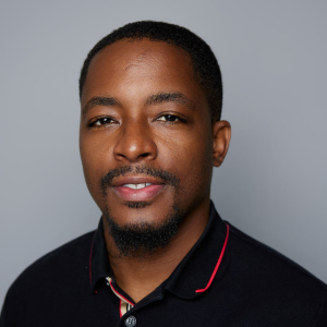 A Black man with short black hair and facial hair is facing the camera and smiling. He is wearing a black shirt with a red logo. The background is gray. - Darius Ballinger