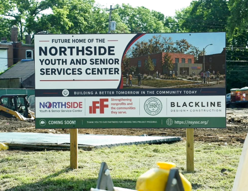 Construction sign for the Northside Youth and Senior Services Center, featuring logos, a construction vehicle, and surrounding buildings and trees.