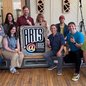 A group of ten people smiling and holding cupcakes in front of a sign that reads "ARTS @ LARGE," indoors with wooden floors and bright lighting.