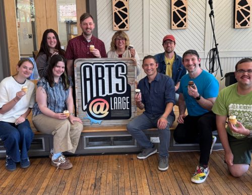 A group of ten people smiling and holding cupcakes in front of a sign that reads "ARTS @ LARGE," indoors with wooden floors and bright lighting.