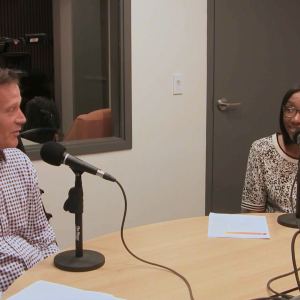 Two people sit at a desk in front of microphones smiling at each other
