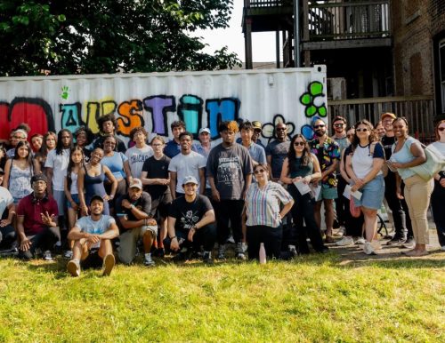 A large, diverse group of people poses outdoors in front of a graffiti-covered shipping container, with "I Love Austin" prominently displayed.