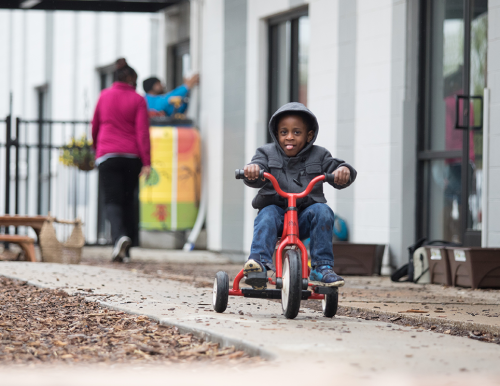 Child in a gray hooded jacket riding a red tricycle along a walkway.