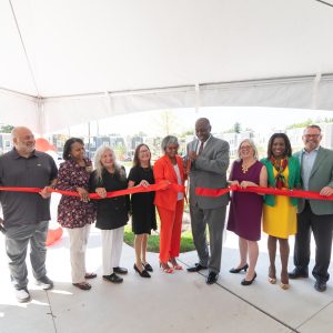 Eleven individuals, dressed in business casual attire, celebrate as Mayor Woods cuts a red ribbon with giant scissors.