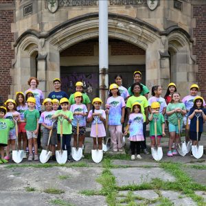 Three rows of children holding shovels and wearing yellow hardhats stand in front of an old brick building's doors.